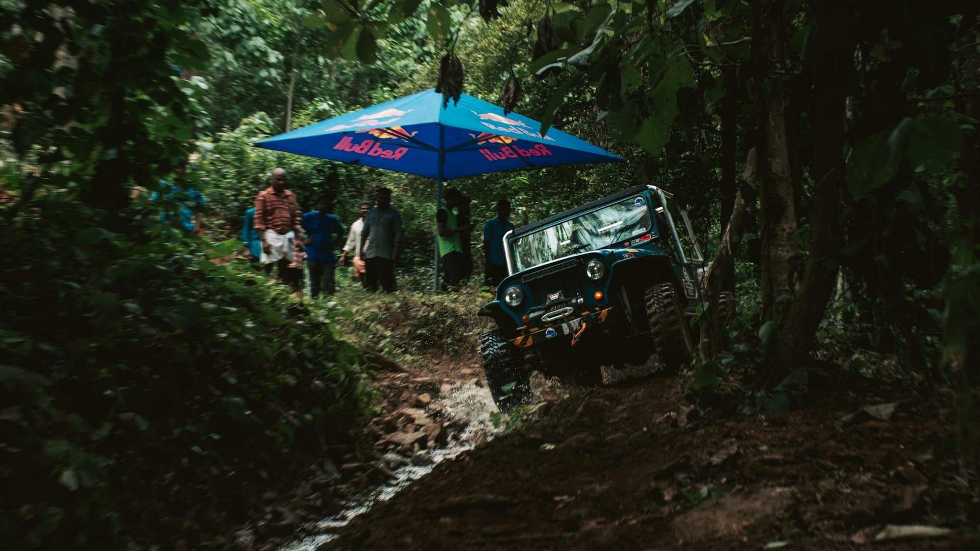 A 4WD off-road jeep climbing through a muddy track in dense tropical jungle, with onlookers and a sun umbrella visible in the background
