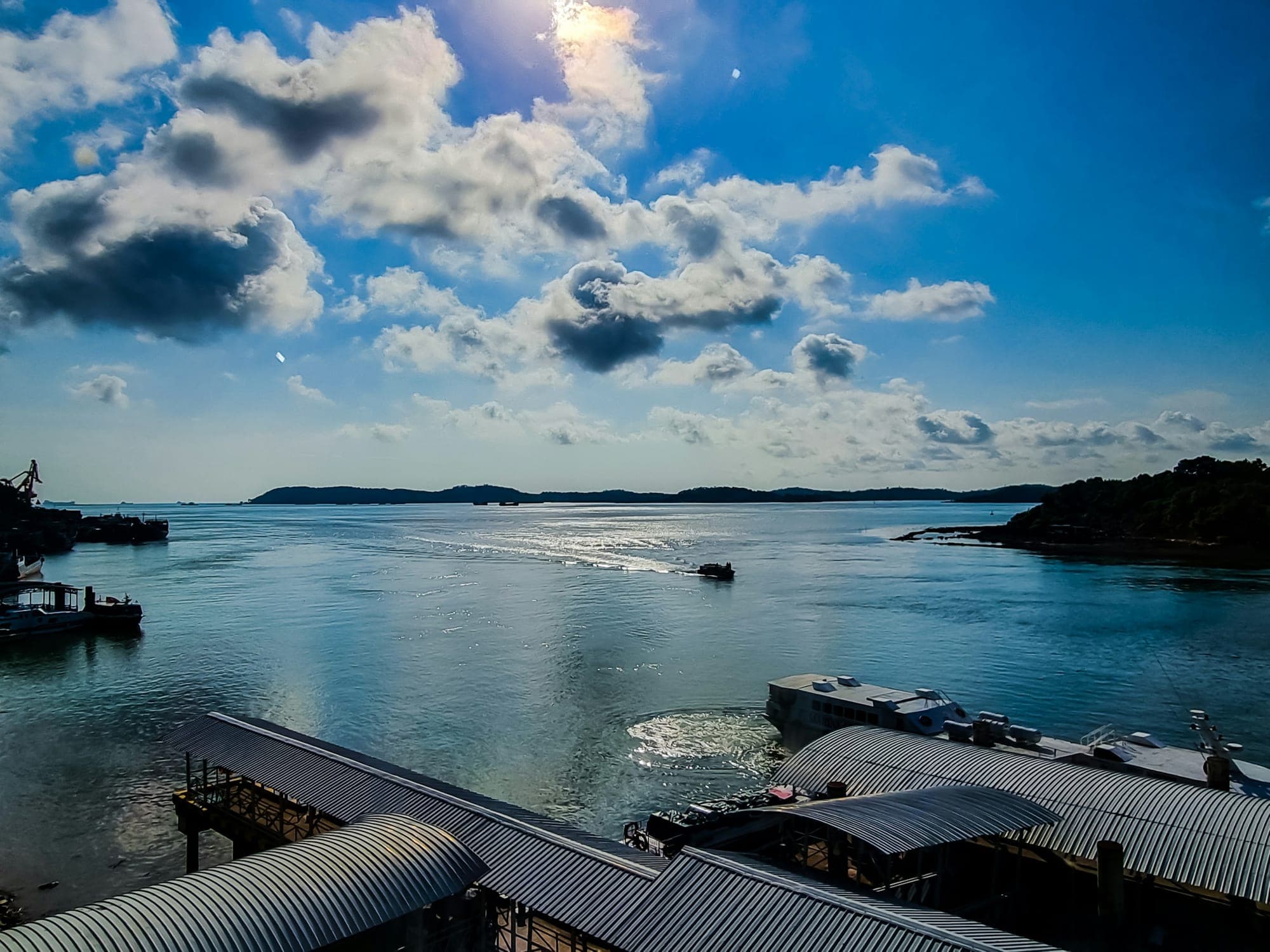 Boats, jetties and corrugated-roof buildings along a harbour on the Batam coast, with islands on the horizon