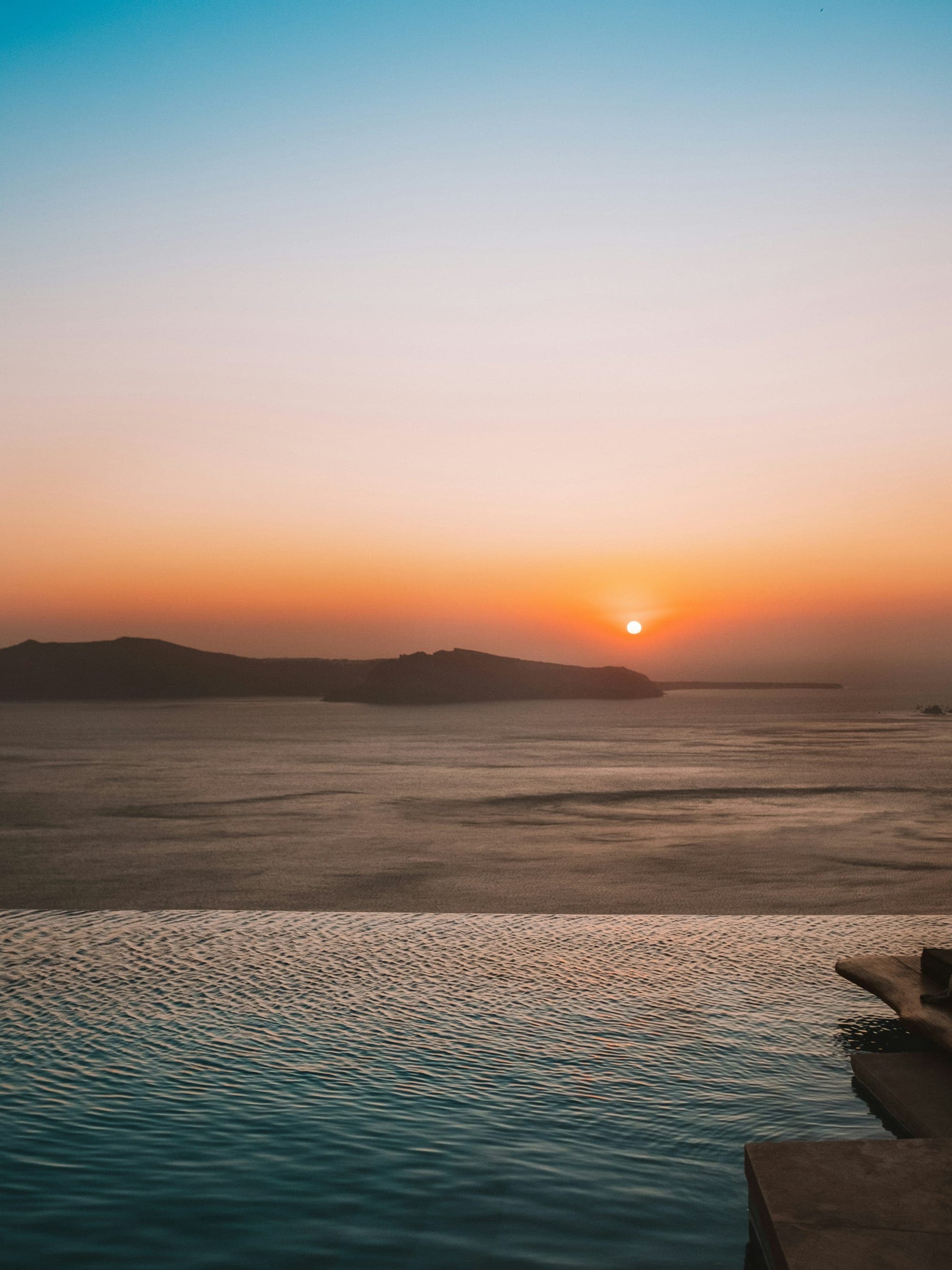 Infinity pool overlooking the ocean at golden hour, with the sun setting behind a distant island silhouette