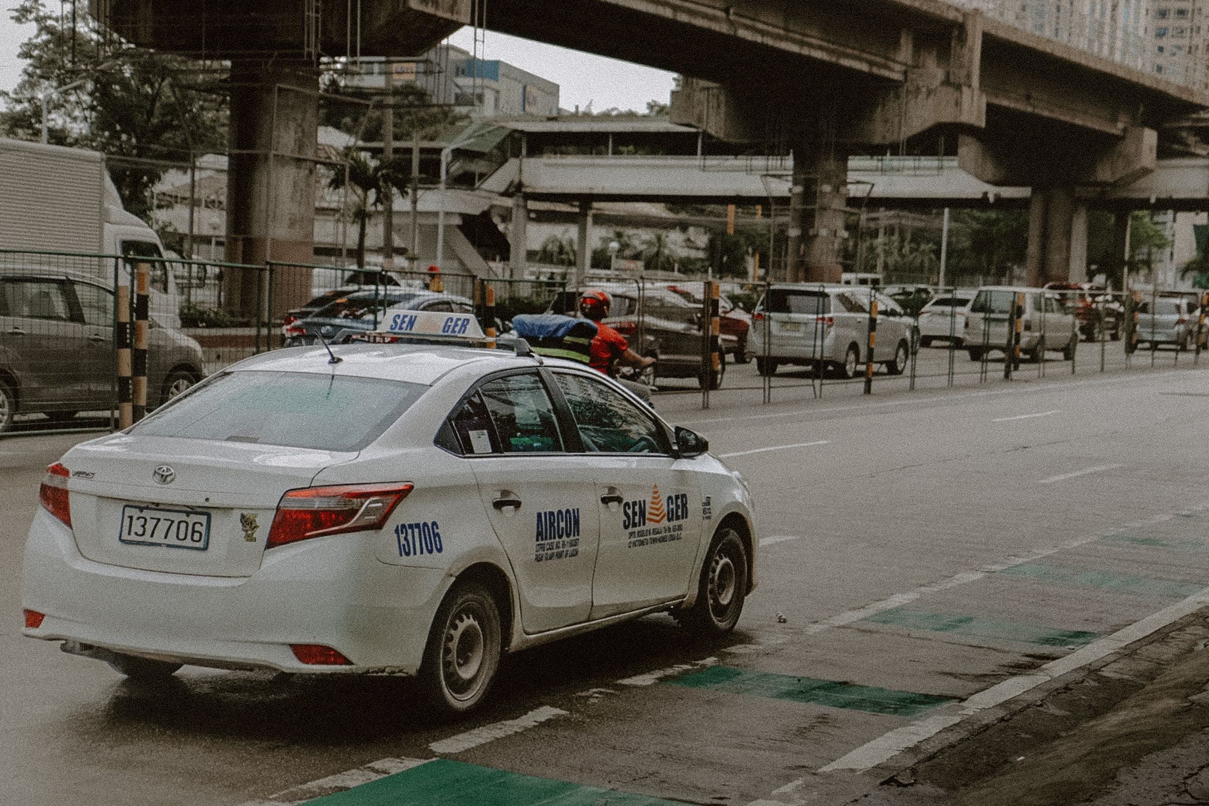 White taxi navigating a busy multi-lane street with overpasses in the background