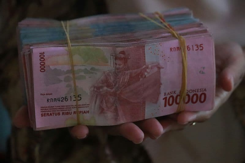 A hand holding a stack of Indonesian Rupiah 100,000 banknotes bundled with a rubber band — the highest-denomination currency note used in Batam