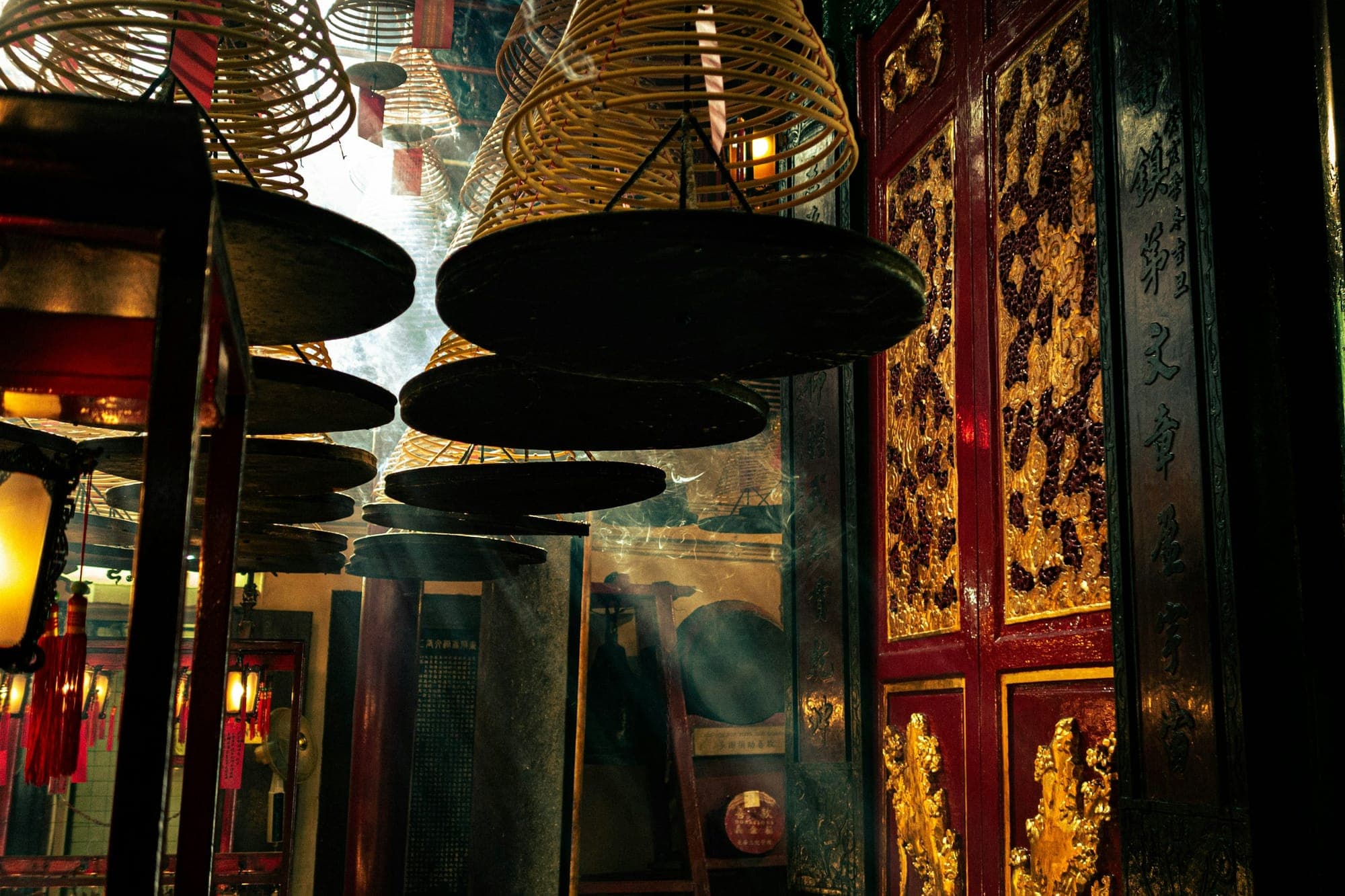 Hanging incense coils suspended from the ceiling of a Chinese-Buddhist temple interior, with red lanterns and gold-carved doorways in warm low light