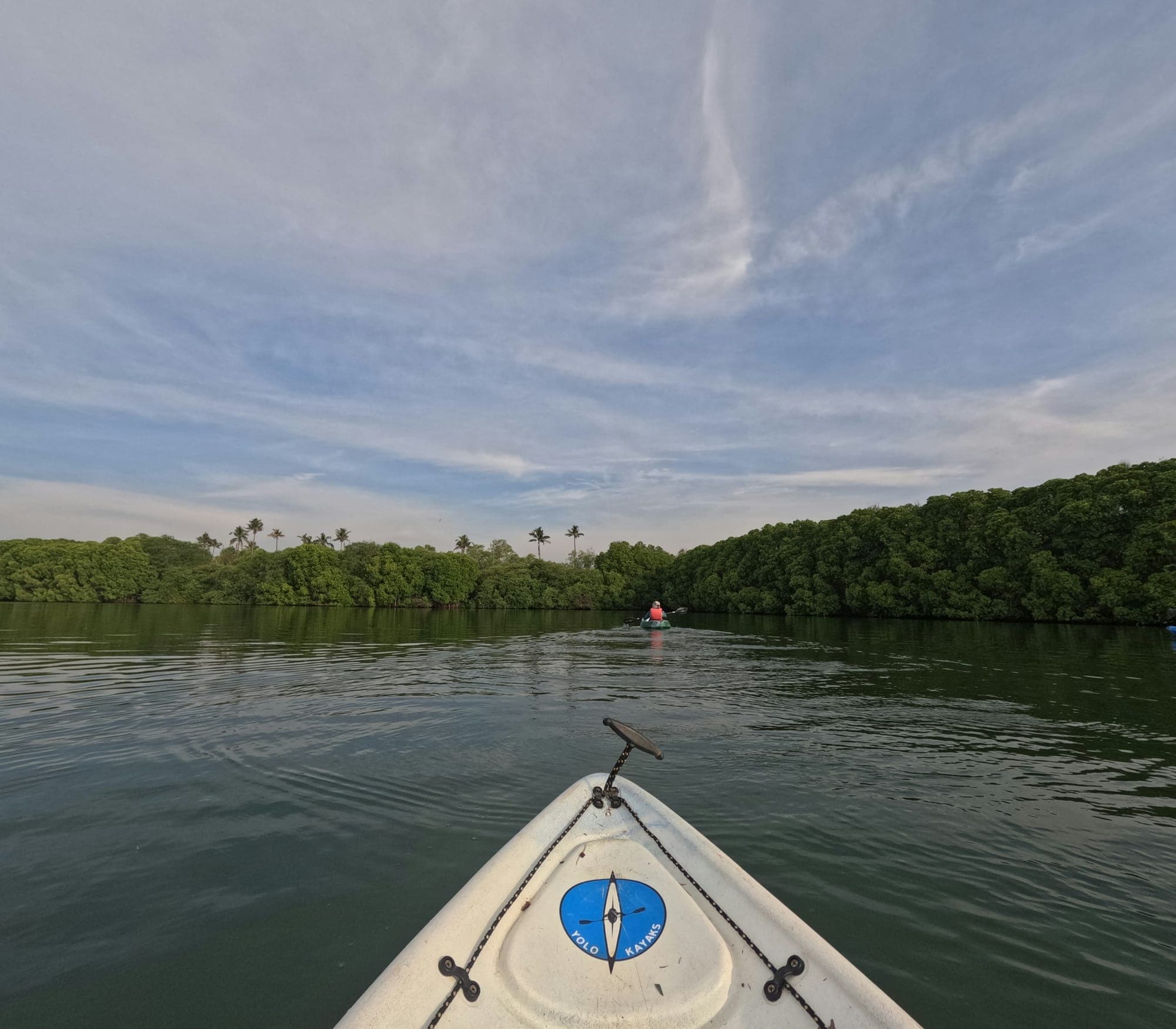 Bow of a kayak gliding through open water with mangrove-lined banks on both sides and a distant paddler visible against a blue tropical sky