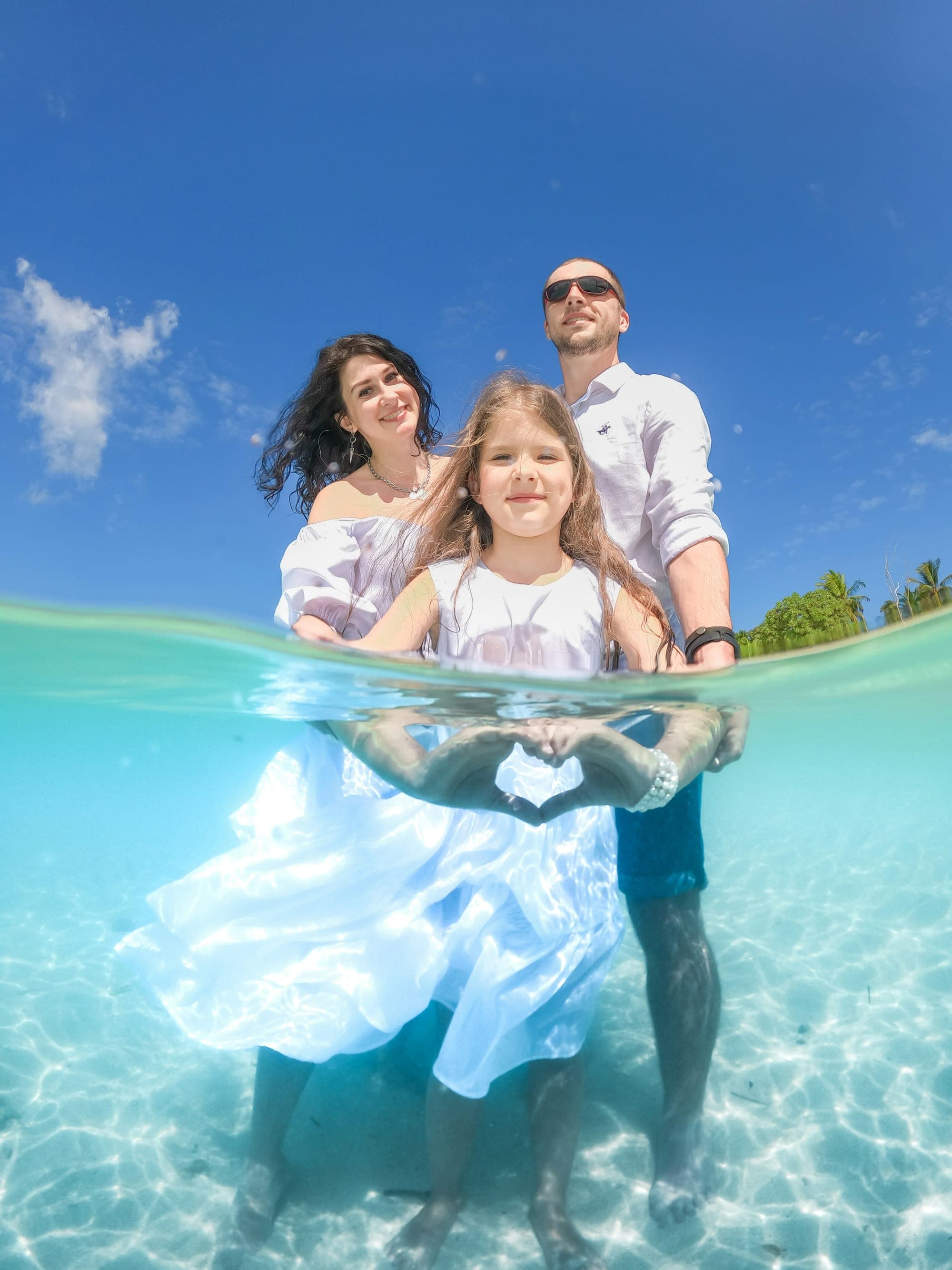 Split-level underwater/above-water shot of a family standing in clear turquoise tropical sea — parents and a young daughter, white sky and palm-trimmed shore behind them