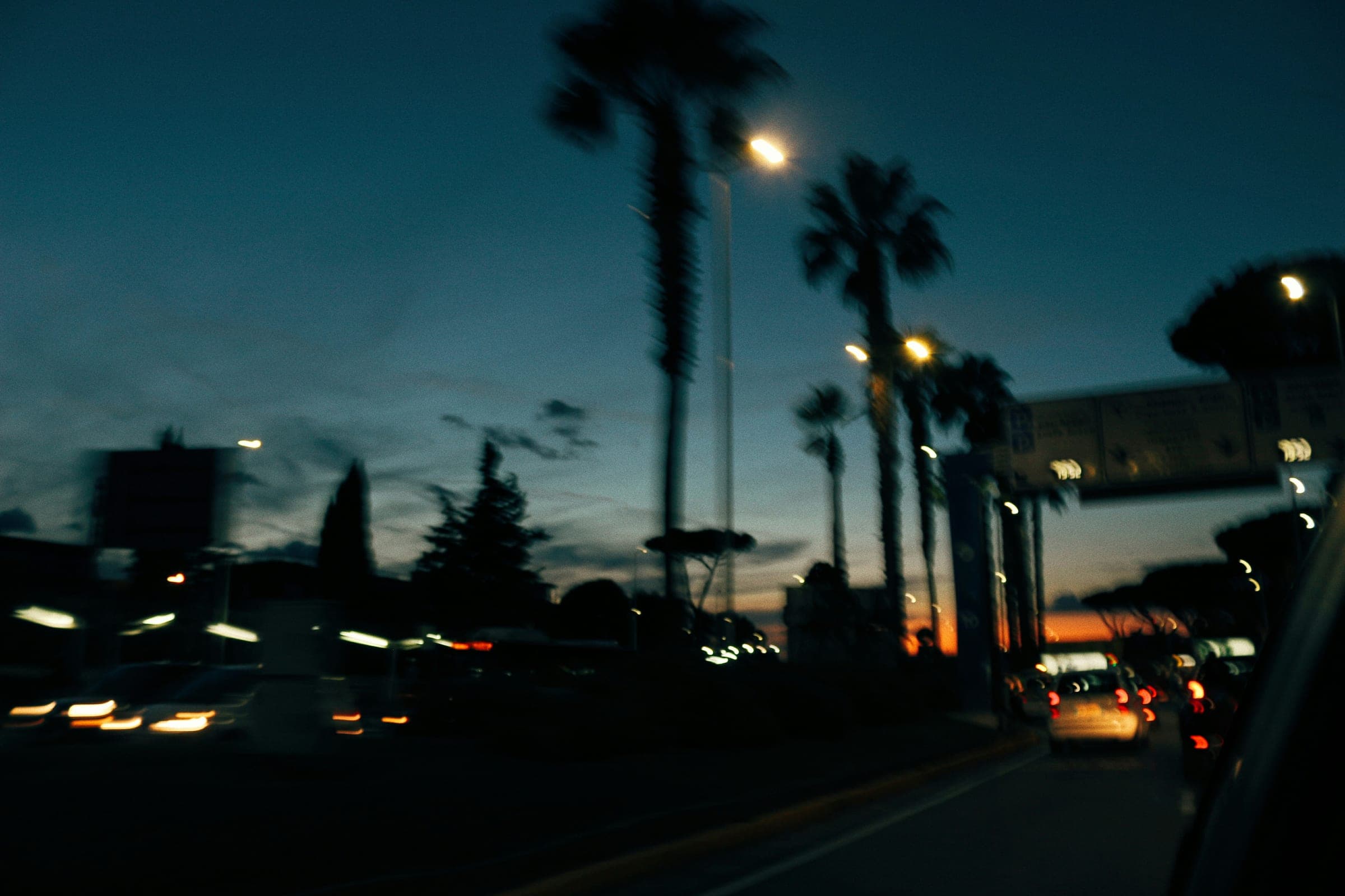 Car driving down a palm-tree-lined street at dusk with the moon visible